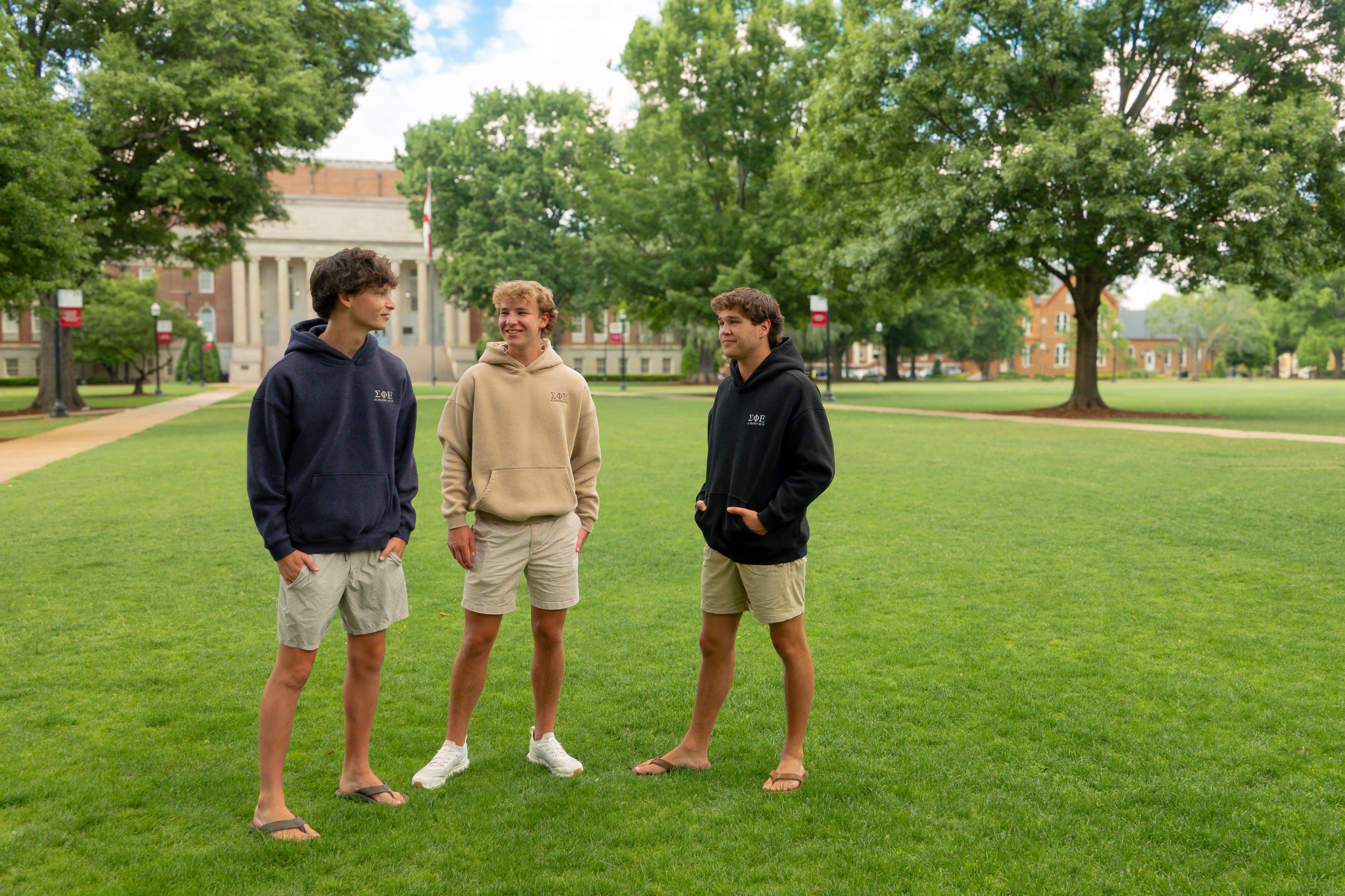 Three young men standing on a grassy field with trees and buildings in the background. On a college campus talking with eachother.