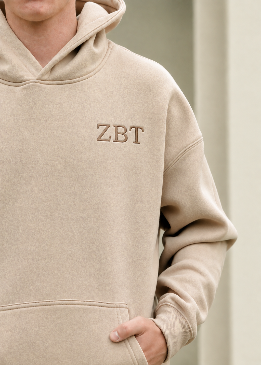 A close-up of a person wearing a beige hooded sweatshirt with the letters 'ZBT' embroidered on the chest, set against a soft neutral background.