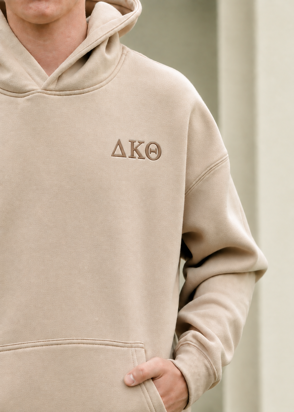 A close-up of a person wearing a beige hooded sweatshirt with the Greek letters Delta Kappa Theta embroidered on the left chest in brown thread.