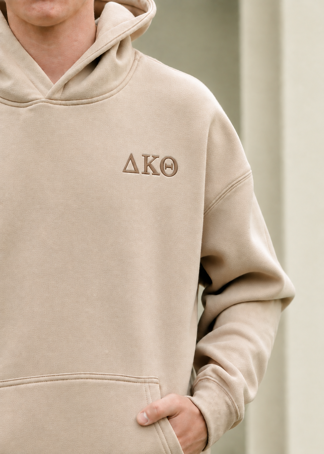 A close-up of a person wearing a beige hooded sweatshirt with the Greek letters Delta Kappa Theta embroidered on the left chest in brown thread.
