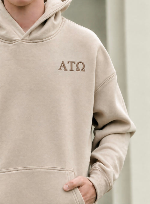 A close-up of a person wearing a beige hooded sweatshirt with the Greek letters Alpha Tau Omega embroidered on the left chest in brown thread.
