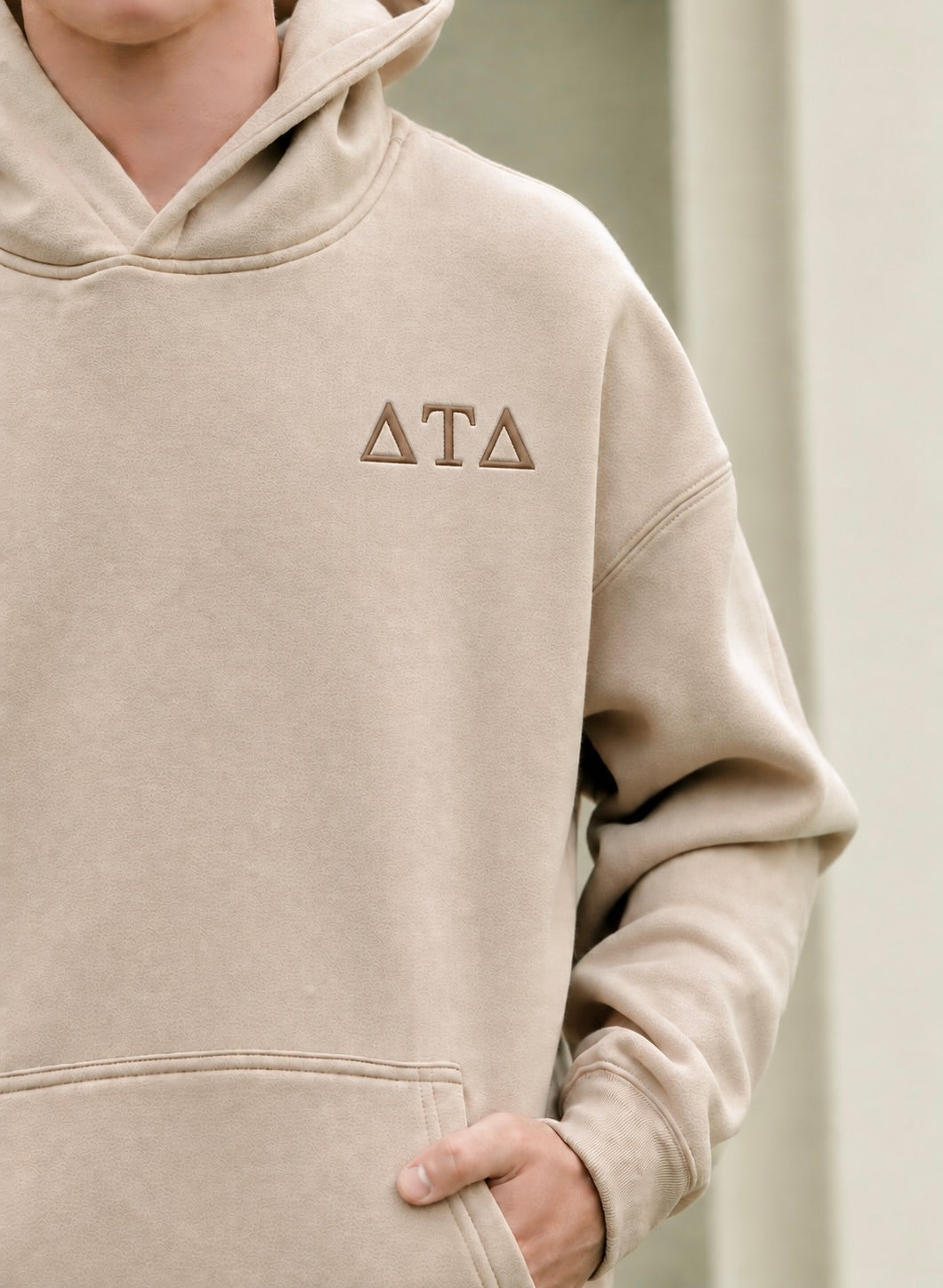 A close-up of a person wearing a beige hooded sweatshirt with the Greek letters Delta Tau Delta embroidered on the left chest in brown thread.