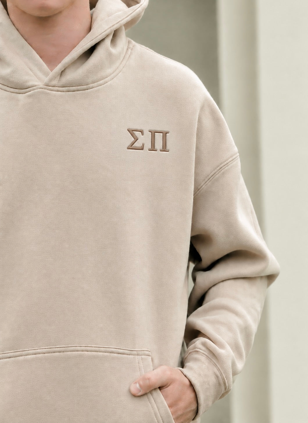 A close-up of a person wearing a beige hooded sweatshirt with the Greek letters Sigma Pi embroidered on the left chest in brown thread.