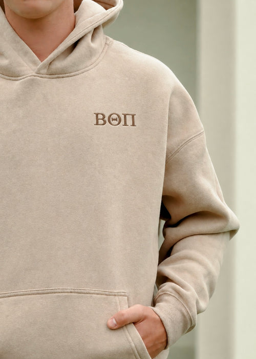 A close-up of a person wearing a beige hooded sweatshirt with the Greek letters Beta Theta Pi embroidered on the left chest in brown thread.