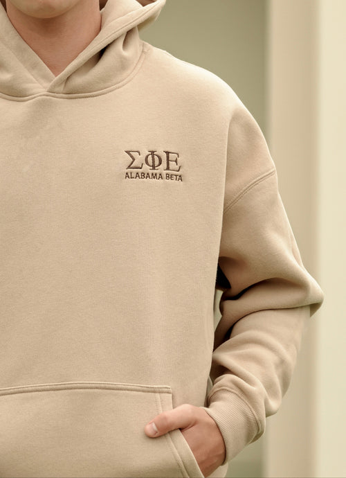 Teenager boy wearing a beige hoodie with embroidered text, Sigma Phi Epsilon Alabama Beta, on a neutral background