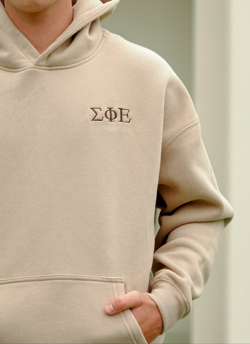 Beige hoodie with embroidered logo of Sigma Phi Epsilon worn by a person. Hand in sweatshirt pocket on a neutral background